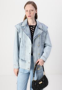 Denim jakna svijetloplave boje s vertikalnim naborima i zlatnim gumbima, uparena s crnom majicom i svijetloplavim trapericama, drži crnu torbicu s kviltanom obradom.