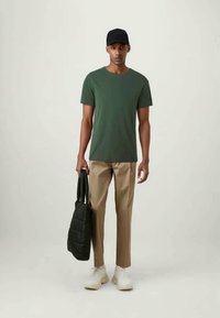Green cotton t-shirt, beige trousers, white sneakers, and a black cap. Holding a black padded tote bag. Simple, casual outfit.