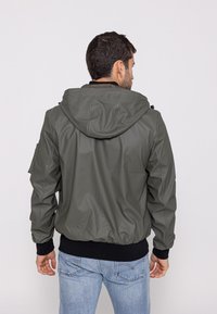 Olive green hooded bomber jacket with a smooth texture, ribbed cuffs, and hem. Features a side pocket on the left arm.