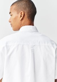 Chemise blanche à manches courtes avec col boutonné, dotée d'un pli au dos et d'une texture en coton lisse. Design simple, sans motifs ni accents.
