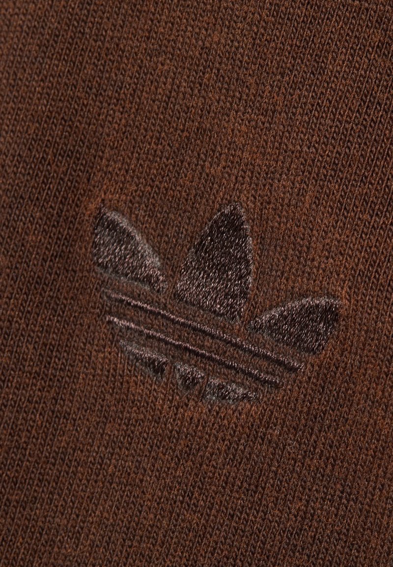 Brauner Strickstoff mit einem dunkelbraunen, gestickten Adidas-Trefoil-Logo in der Mitte des Materials.