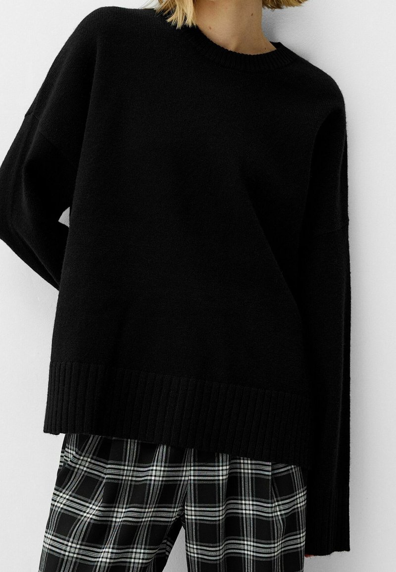 Personne portant un pull noir ample avec ourlet et poignets côtelés, associé à un pantalon à carreaux noir et blanc, sur un fond blanc uni.