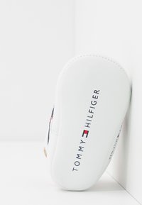 Zapatilla blanca con punta redonda, con parte superior de cuero texturizado, logo bordado y detalles en azul marino y rojo.