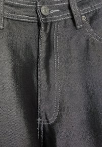 Črne jeans hlače iz denima z vidnimi šivi, kovinsko zaponko in sprednjimi žepi. Tkanina ima rahlo sijaj in teksturiran zaključek.