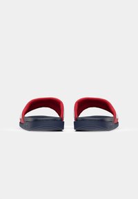 Røde og marineblå slidesandaler med en blød, tekstureret rem og en polstret sål, der har en buet form og et minimalistisk design.