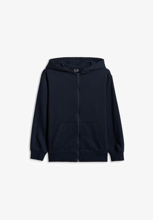 Enfant debout portant un bonnet rouge, un sweat à capuche bleu marine zippé, un pantalon cargo gris et des baskets grises avec des lacets roses sur un fond uni.