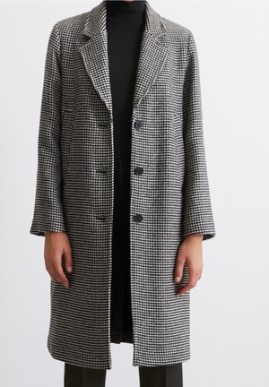 Sort og hvid hundetandsmønstret lang frakke med revers og tre knapper båret over en sort rullekrave og bukser.