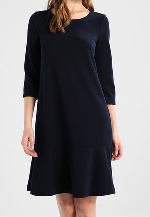 Frau trägt ein schlichtes marineblaues, knielanges Kleid mit dreiviertel Ärmeln und ausgestelltem Saum vor einfarbigem Hintergrund.