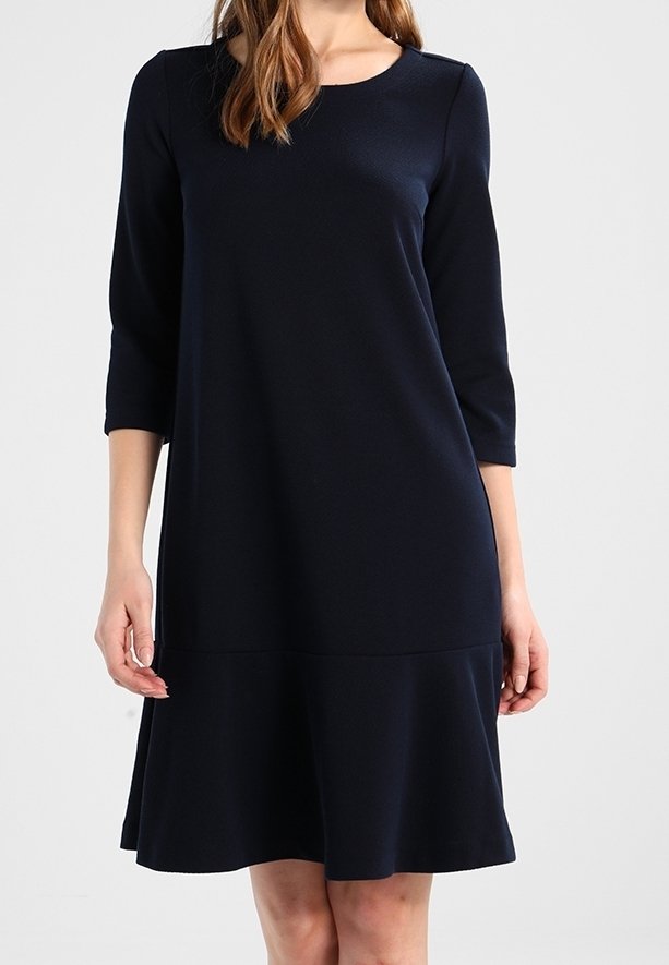 Femme portant une robe simple bleu marine, longueur genou, manches trois-quarts et ourlet évasé, sur un fond uni.