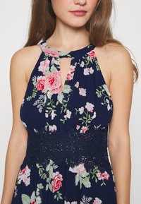Kvinna klädd i en ärmlös marinblå klänning med rosa och vita blommönster och ett brett midjeband i marinblå spets, som visar axlar och övre bröst.