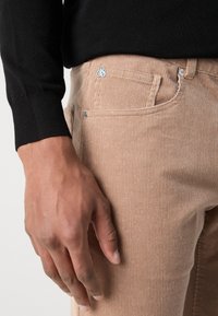 Pantaloni bej din corduroy cu dungi verticale, având un logo albastru deschis pe brâul de la talie. Un pulover negru este parțial vizibil pe braț.