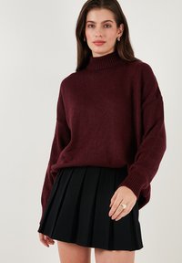 Bordó pulóver magas nyakkal és leeresztett vállakkal, fekete plisszírozott mini szoknyával párosítva. Sima textúra bordázott mandzsettákkal.