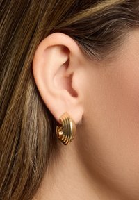 Boucles d'oreilles en or avec un design strié et des bords lisses et incurvés, mises en valeur contre un gros plan d'une oreille et de cheveux.