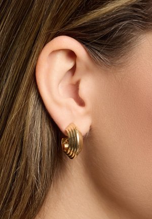 Goldene Creolen mit einem gerippten Design und glatten, abgerundeten Kanten, die vor einer Nahaufnahme eines Ohres und Haaren präsentiert werden.