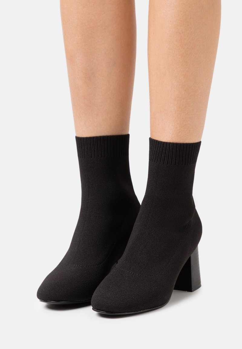 Bottes chaussettes en maille noire avec talons blocs portées sur des jambes nues, sur un fond neutre.
