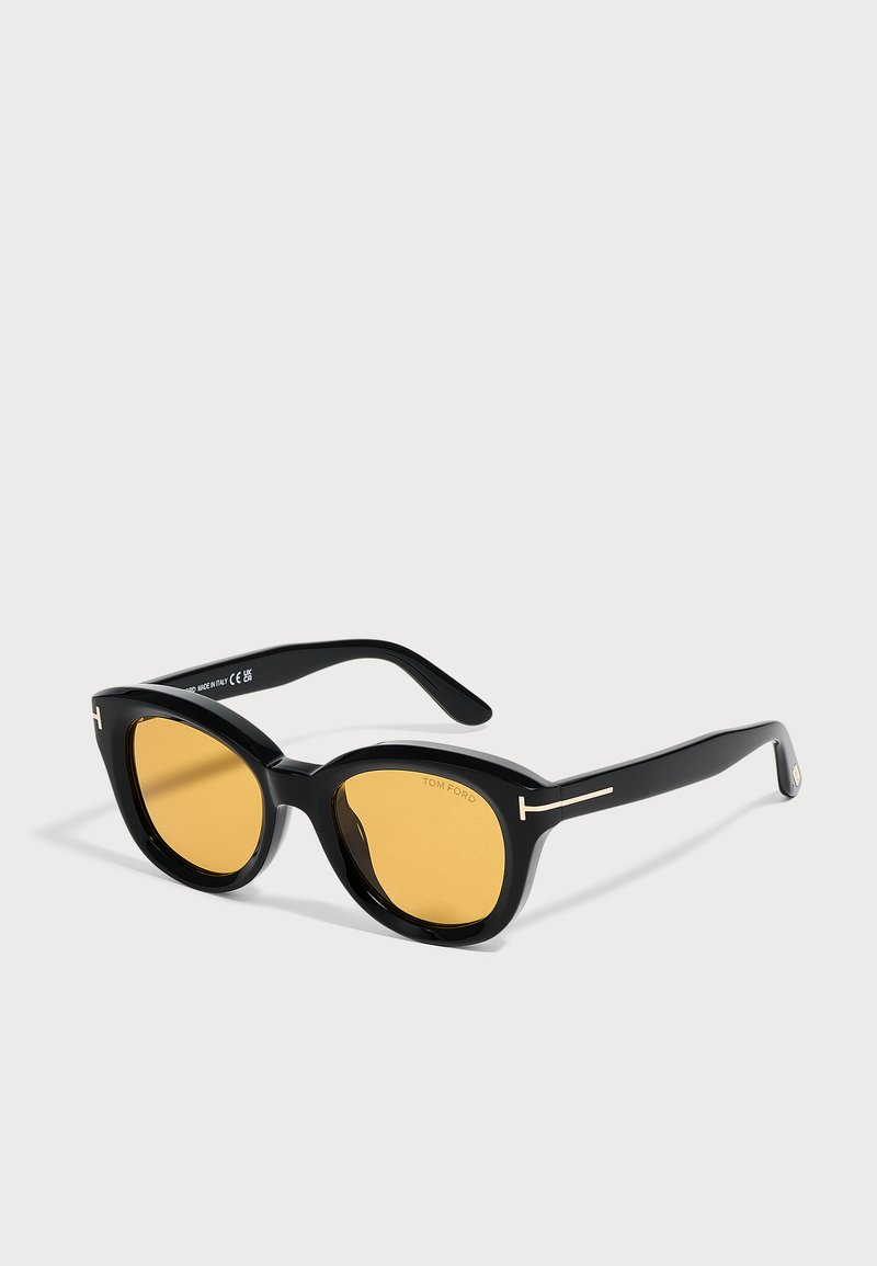 Lunettes de soleil noires à monture ronde avec verres jaunes et accents dorés en forme de T sur les branches, présentées sur un fond neutre.