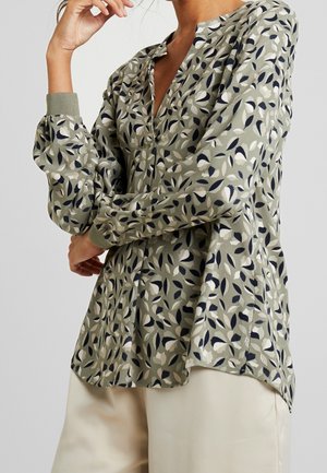 Femme portant un chemisier vert à manches longues avec un motif de feuilles noir et blanc, associé à un pantalon beige clair.