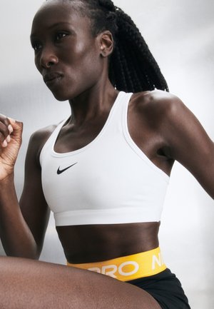 Mujer atlética con sujetador deportivo blanco y pantalones cortos negros con cintura amarilla, concentrada y corriendo o haciendo ejercicio en interiores con fondo neutro.