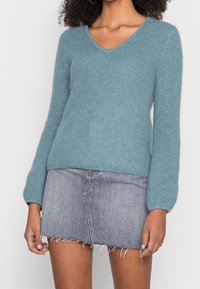 Türkisfarbener Strickpullover mit V-Ausschnitt und langen Ärmeln. Weicher Griff und figurbetonte Silhouette, kombiniert mit einem grauen Jeans-Minirock mit ausgefranstem Saum.