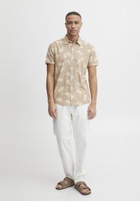 Kortermet, beige knappskjorte med et kremfarget sirkulært mønster, parret med hvite bukser og brune sandaler. Enkelt og uformelt design.