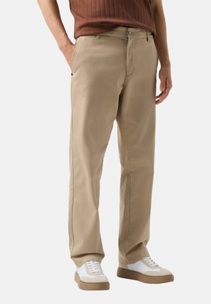 Mand iført beige bukser med lige ben og hvide sneakers, stående med den ene hånd i lommen mod en ensfarvet baggrund.