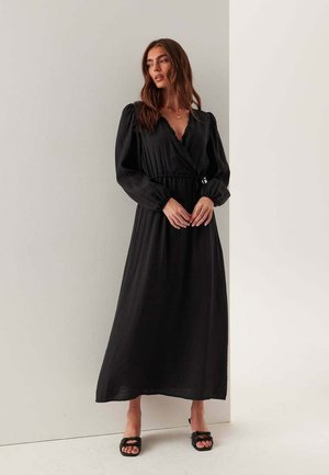 Donna con capelli castani lunghi che indossa un vestito nero a portafoglio a maniche lunghe e sandali neri con tacco, in piedi contro un muro bianco.