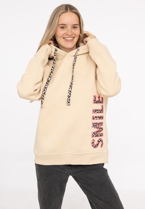 Haut à capuche crème avec un accent en imprimé léopard sur les cordons de serrage et le mot "SOURIRE" en lettres audacieuses et à motif sur le côté.