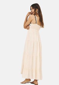 Vestido maxi de tejido beige claro, con tirantes ajustables, dobladillo con volante y cuerpo fruncido con acentos negros en forma de escallopado.