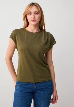 Blondynka w oliwkowozielonej bluzce z krótkim rękawem i niebieskich dżinsach, stojąca na jasnym, jednolitym tle, z jedną ręką w kieszeni.