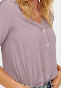 Blusa lilla con scollatura a V, maniche corte e cinque bottoni sul davanti. Tessuto morbido con una leggera texture, abbinata a jeans a vita alta.