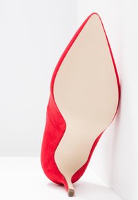 Chaussure à talon haut en suède rouge à bout pointu, vue de la semelle, debout contre un fond blanc.