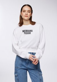 Felpa bianca cropped con stampa nera "NO DRAMA PLEASE" sul davanti, abbinata a pantaloni cargo in denim con tasche laterali.