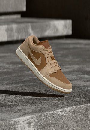 Tan- og brun Nike-sneaker i luften over en betonoverflade med trapper i baggrunden.