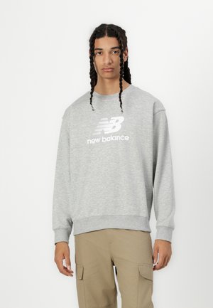 Sudadera gris con cuello redondo, mangas largas y puños acanalados, que presenta un logotipo blanco de "New Balance" en la parte frontal. Combinada con pantalones cargo beige.