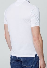 Polo shirt blanc à manches courtes avec col, en tissu texturé et fentes latérales, associé à un jean en denim bleu.