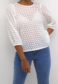 Haut en dentelle blanche avec un motif travaillé et des manches trois-quarts. Associé à un jean en denim bleu clair, présentant une silhouette ajustée.