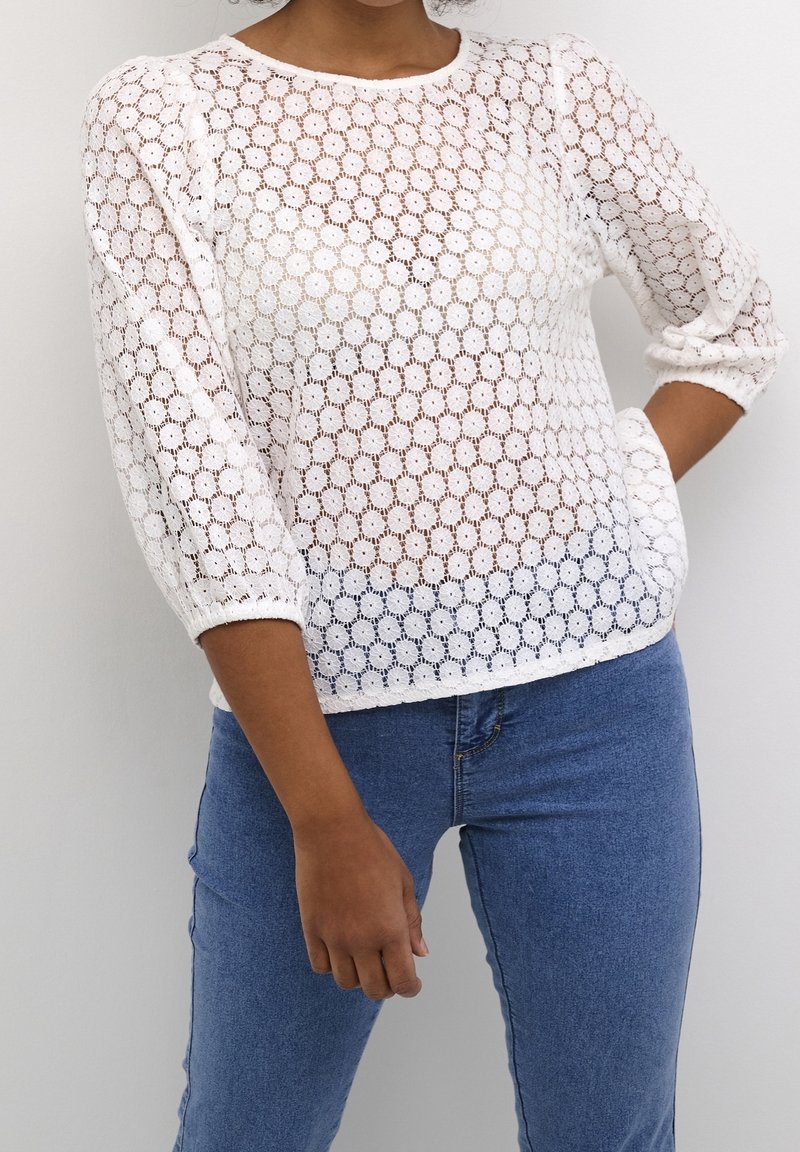 Haut en dentelle blanche avec un motif travaillé et des manches trois-quarts. Associé à un jean en denim bleu clair, présentant une silhouette ajustée.