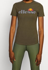Camiseta ajustada verde oliva con un cuello redondo, que presenta un logo gris "ellesse" y un gráfico de sol colorido encima.