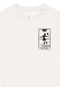 Witte katoenen t-shirt met een kleine zwarte afbeelding van een figuur die op een klif balanceert, gelabeld "DE DWAAS" met een zon erboven.