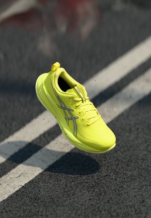 Chaussure de course jaune vif avec des rayures noires et blanches suspendue au-dessus d'une route en asphalte marquée de lignes blanches.
