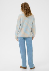 Cilvēks ar cirtainiem matiem, tērpies bēšā un gaiši zilā rakstainā džemperī, gaiši zilās džinsās un brūnās kurpēs, stāv ar muguru pret baltu fona.