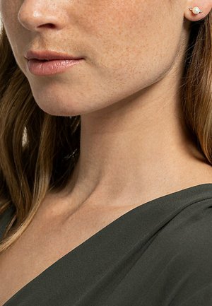 Goldene Ohrringe mit einer kleinen Perle, strukturierte Haut mit Sommersprossen und einem dunkelgrünen Kleidungsstück. Nahaufnahme von Hals und Gesicht.