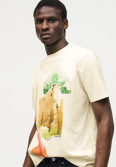 Άνδρας που φοράει κρεμ μπλουζάκι με πολύχρωμο σχέδιο αρχαίων αιγυπτιακών αγαλμάτων και ερειπίων, στέκεται μπροστά σε λευκό φωτεινό φόντο.