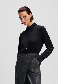 Camicia nera con bottoni, caratterizzata da una finitura lucida, colletto a punta e maniche lunghe. Abbinata a pantaloni grigi a righe sottili.