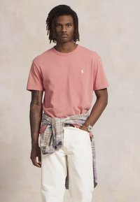 Νεαρός άνδρας με κοτσιδάκια, ντυμένος με ροζ μπλούζα, λευκά παντελόνια, και πουκάμισο καρό δεμένο στη μέση, κρατώντας ρολόι, ενώ στέκεται μπροστά από απλό φόντο.