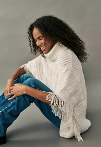 Weiße gestrickte Poncho mit hohem Kragen, der mit texturierten Mustern und fransigen Kanten versehen ist, kombiniert mit blauen Jeans aus Denim, die mit weißen Nieten verziert sind.