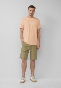 T-shirt di cotone color pesca chiaro con piccolo logo bianco, abbinata a pantaloni corti verde oliva e sneakers crema con suola marrone. Vestibilità e design casual.
