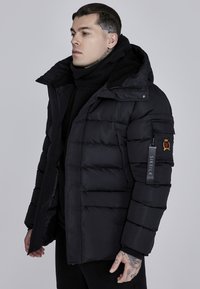 Sort pufferjakke med aftagelig hætte, fremtrædende syninger, sidelommer, branding på ærmet og en blød inderside.