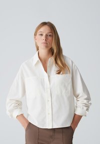 White button-up shirt with two front pockets, featuring a collar and rolled-up sleeves, made of textured fabric.