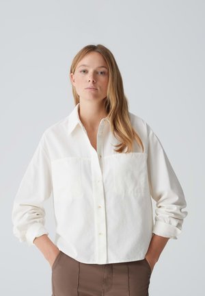 White button-up shirt with two front pockets, featuring a collar and rolled-up sleeves, made of textured fabric.